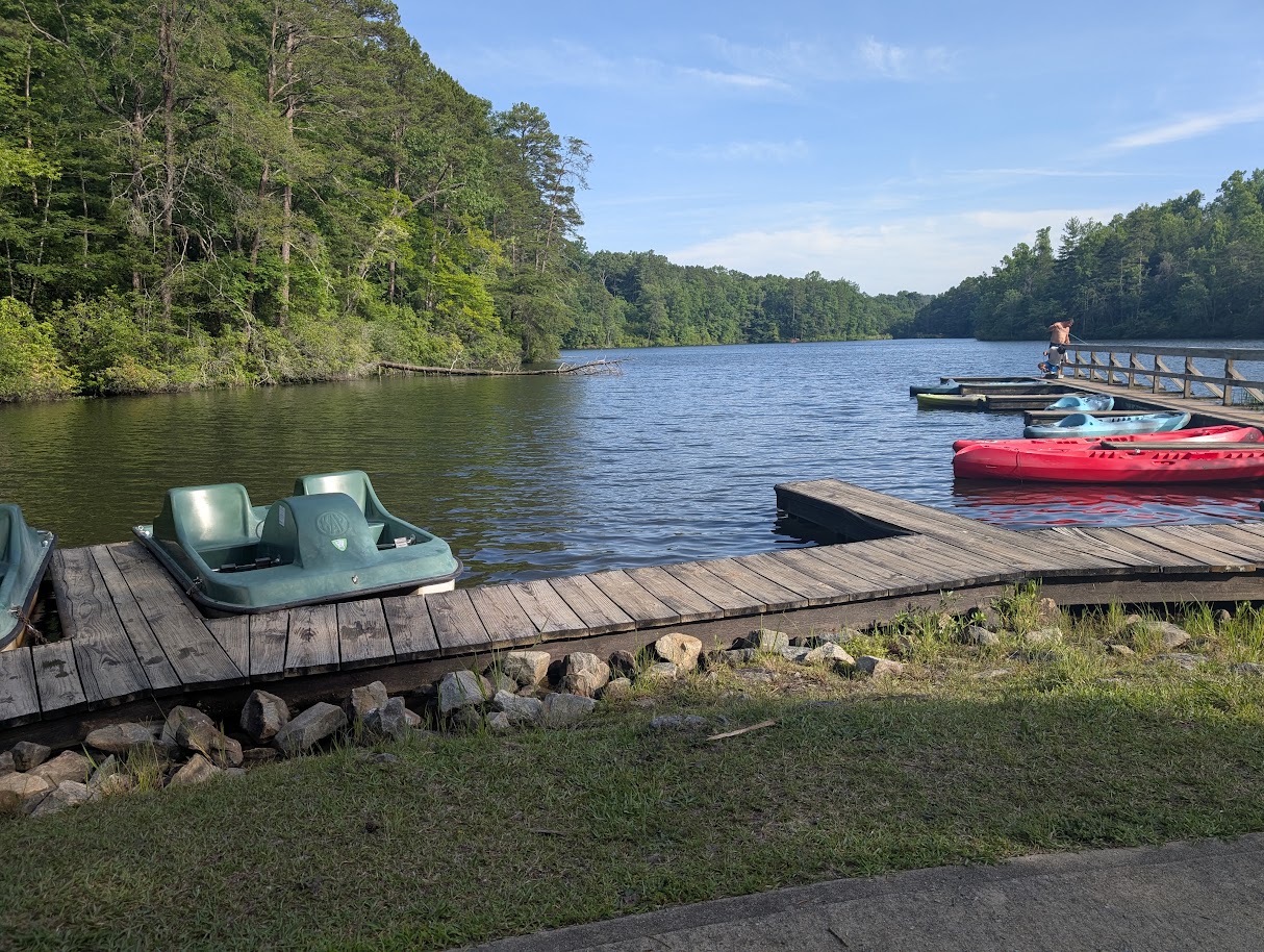 Kayaks and pedalboats at a wooden dock beside a forested lake.