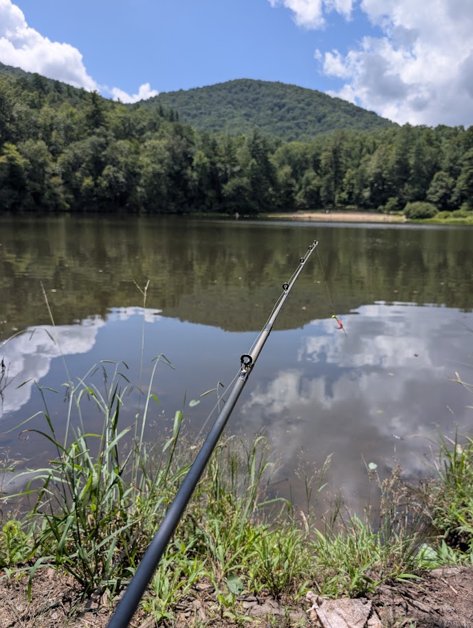A fishing rod beside a calm lake backed by forested hills.